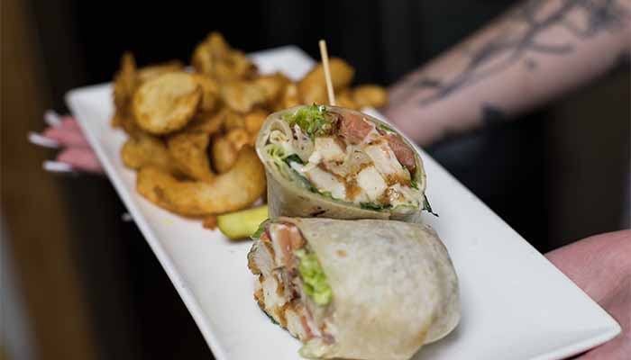 A Cadence Clubhouse chicken wrap cut in half on a plate with a side of potato wedges.